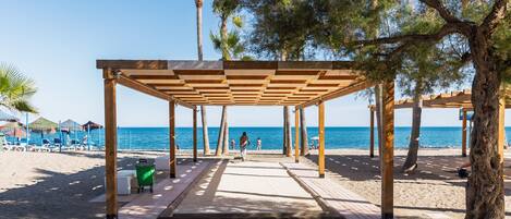 A beachside walkway with wooden pergolas, palm trees, and beach umbrellas.