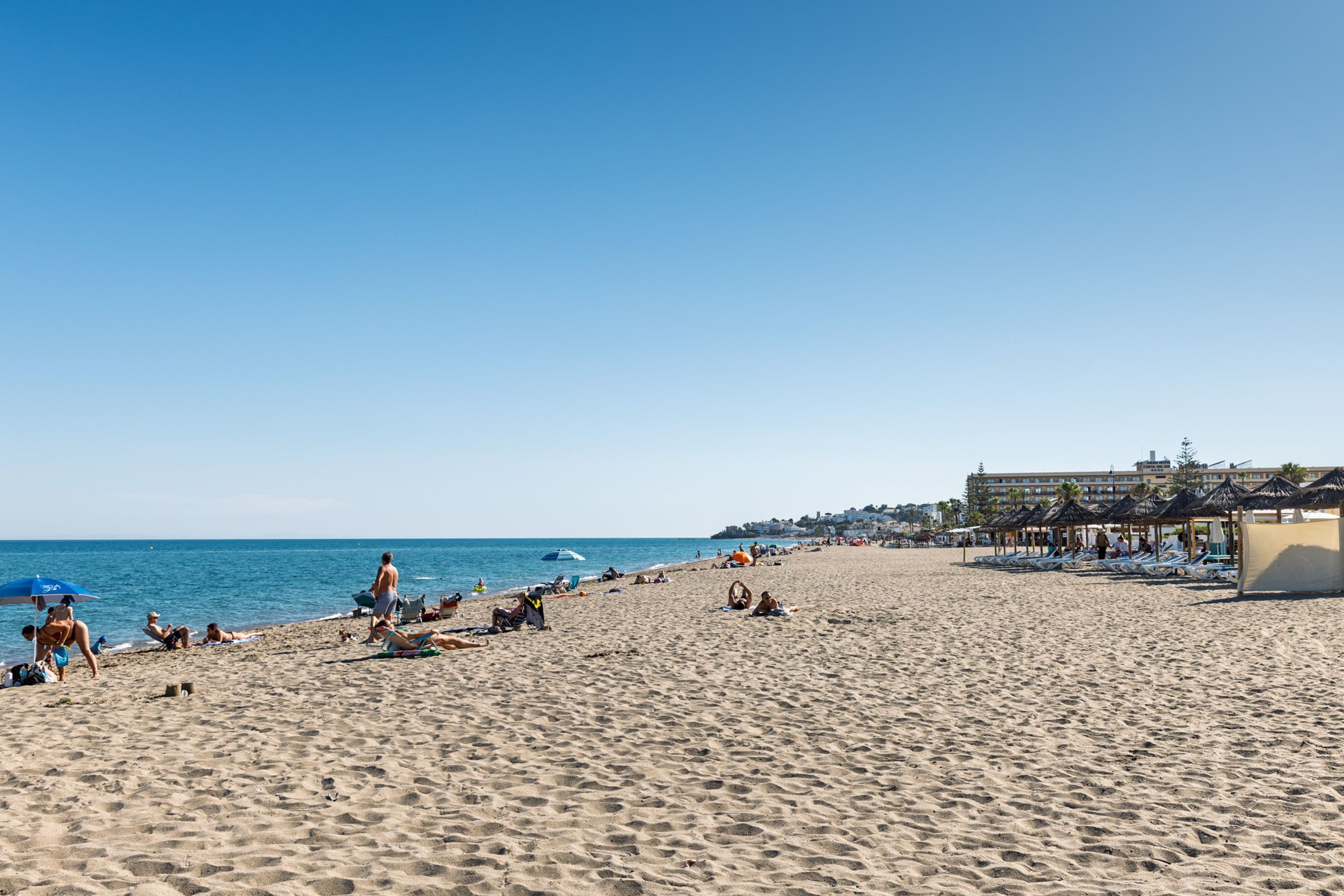 Een strand met mensen die zonnebaden en zwemmen, een gebouw met uitzicht op zee en een heldere lucht.