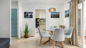 Uma sala de jantar com mesa e cadeiras de vidro, uma planta em vaso e vista para a cozinha.