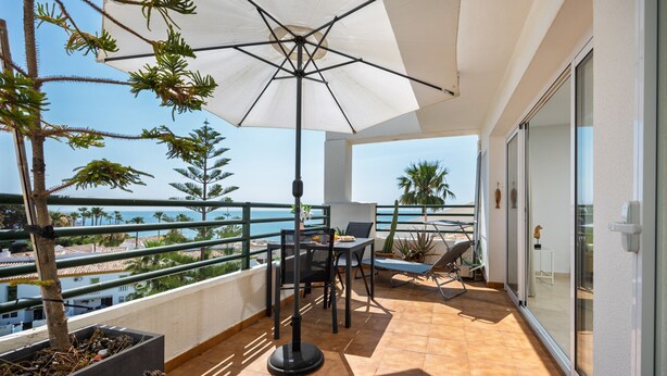 Uma varanda com mesa, cadeiras, um guarda-sol e plantas em vasos.