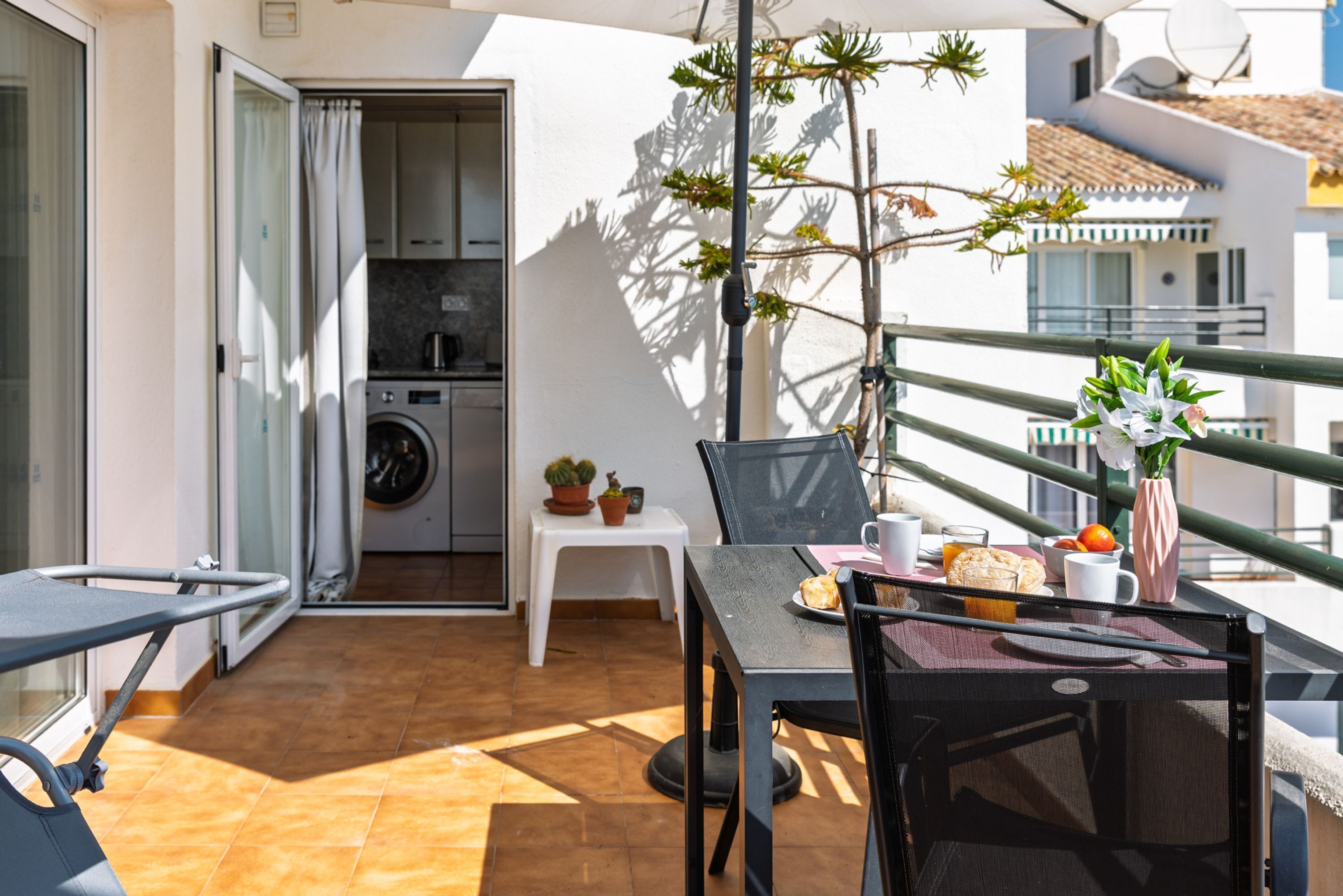 A balcony with a table, chairs, a small table with a plant, and a potted plant.
