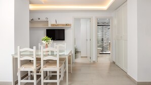 Et moderne spiseområde med et glasbord, hvide stole, et fladskærms-tv og en gang, der fører til en trappe.