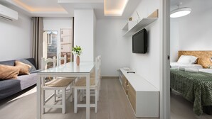Et moderne, lyst værelse med et glasbord, hvide stole, en sofa, en seng med et grønt tæppe, et vægmonteret tv og en lille plante.