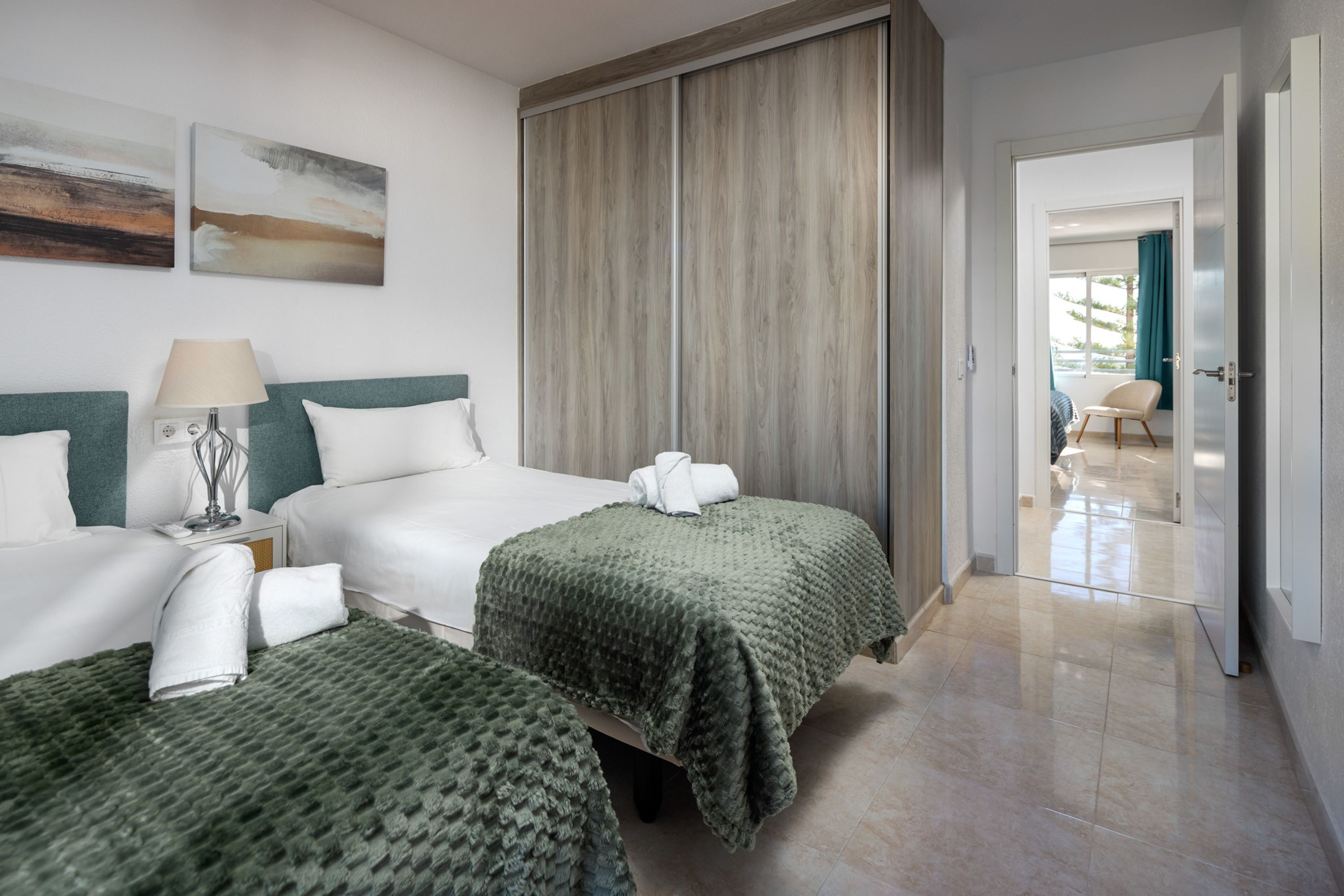 Een hotelkamer met twee bedden, een nachtkastje, een lamp, een grof geweven groene deken en een houten garderobe.