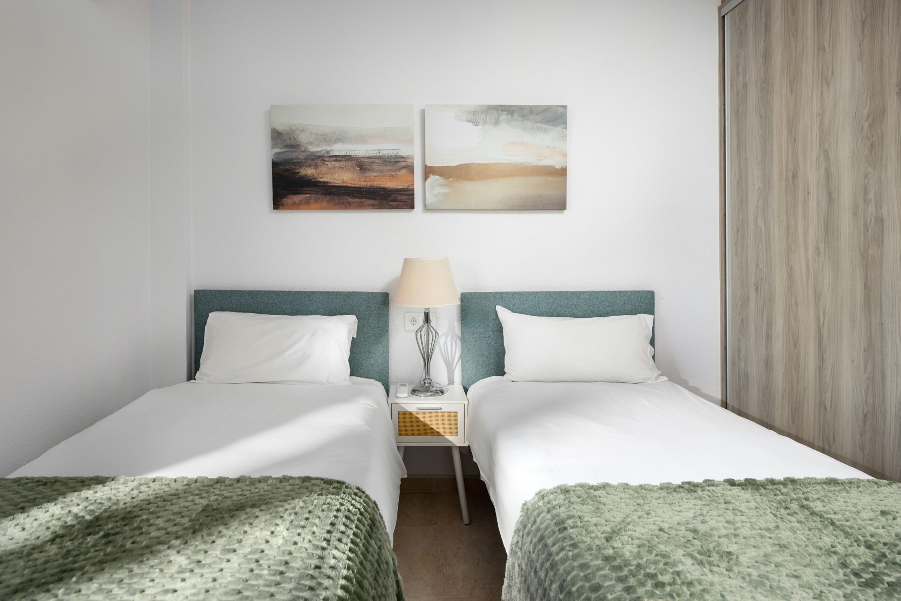 Ein Hotelzimmer mit zwei Betten, einem Nachttisch mit Lampe und zwei abstrakten Bildern an der Wand.