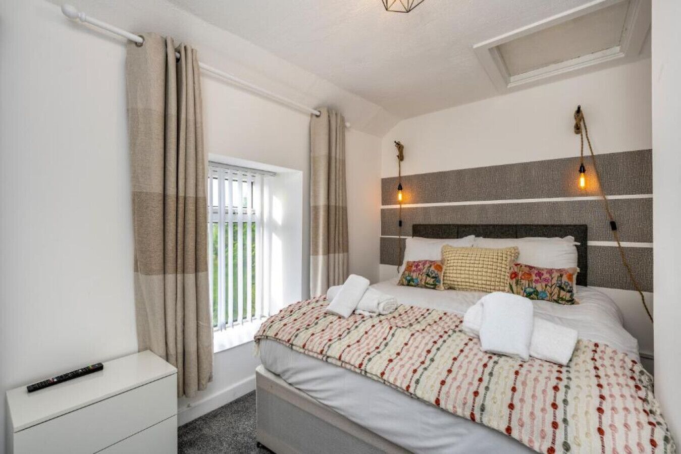Une chambre à coucher avec un lit, des rideaux, une table de chevet, une télécommande et une fenêtre avec des stores.