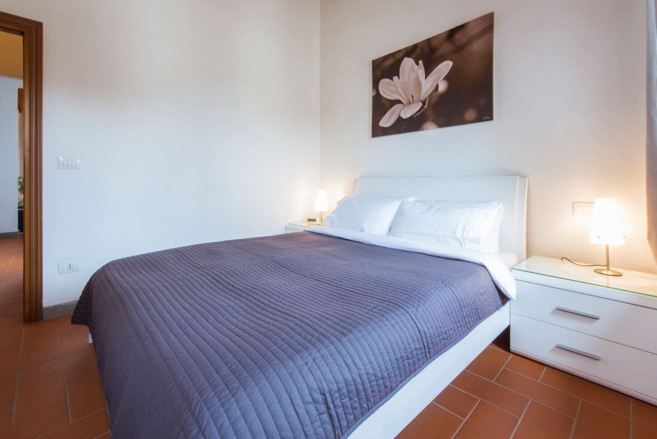 A neatly made bed with a dark quilt, a white headboard, and a bedside table with a lamp.