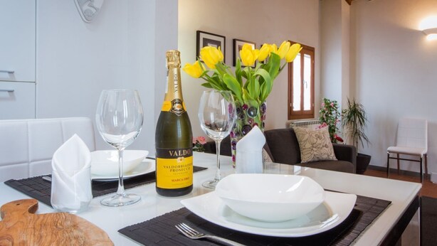 A dining table set with a bottle of Valdo champagne, two wine glasses, a vase with yellow flowers, and a white plate. - Apartments Florence - Boboli Balcony Rooftop View Florence City Center (Firenze)