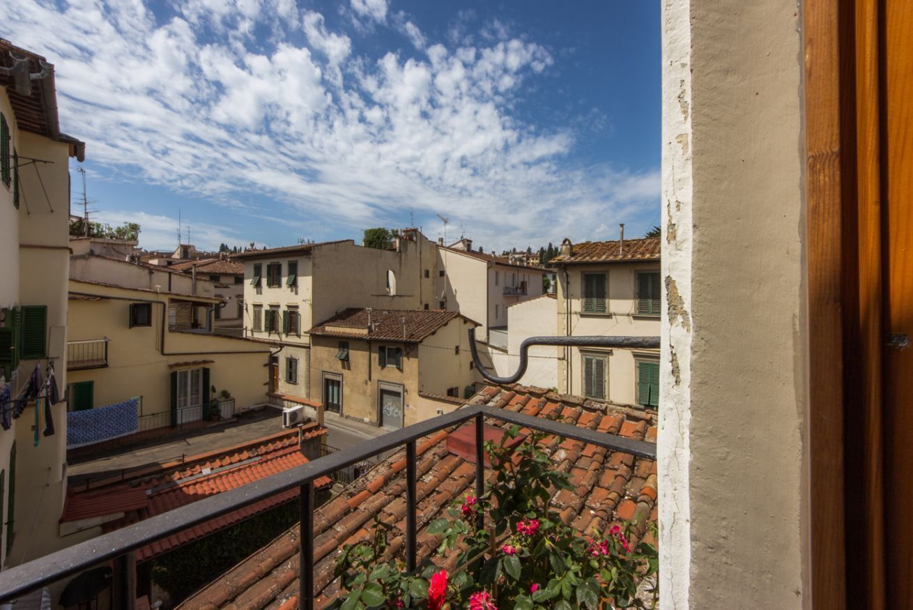 Un balcon donnant sur des bâtiments aux toits de tuiles rouges, avec une balustrade et une jardinière remplie de fleurs roses.