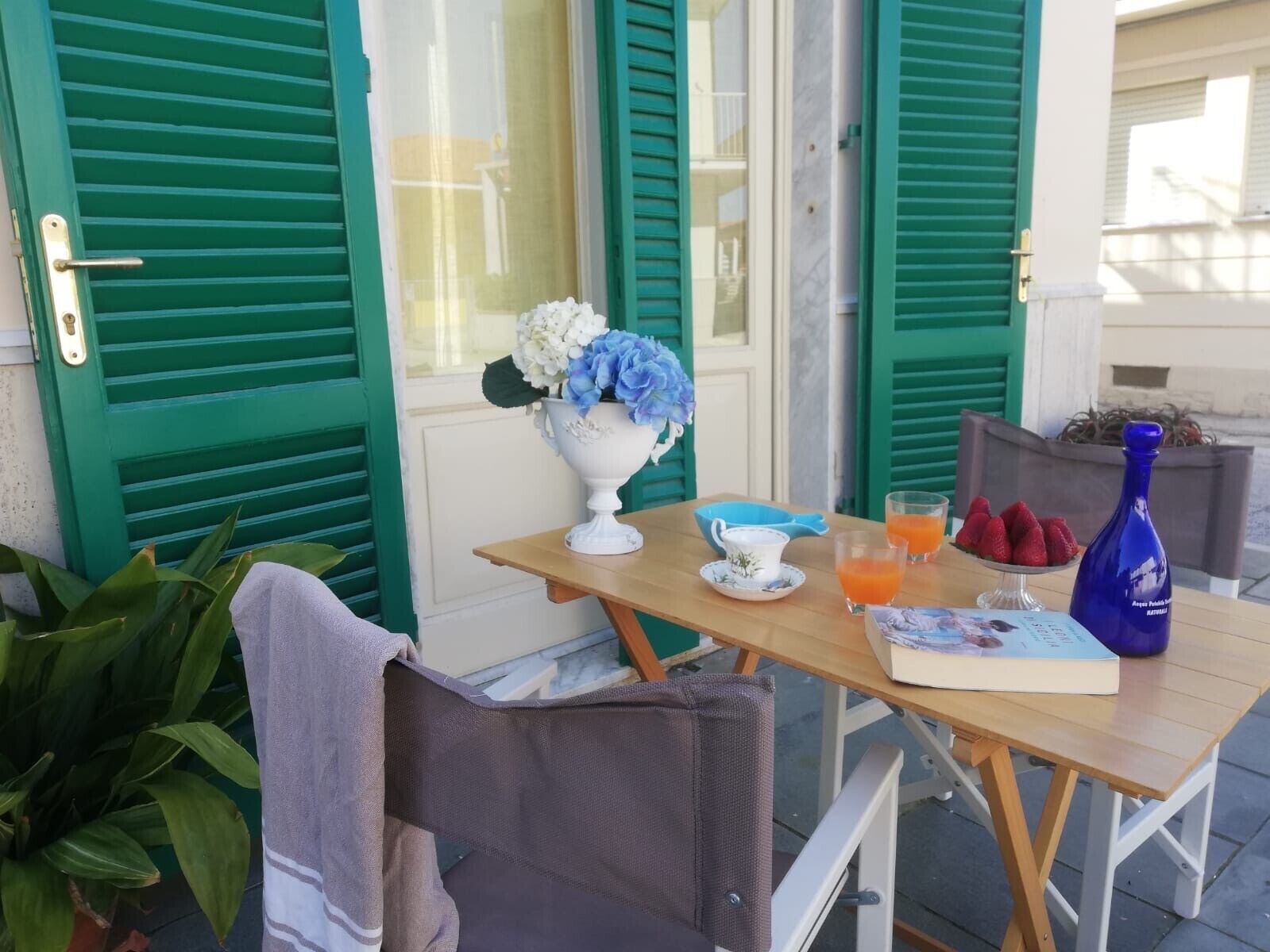A table set outdoors with a book, a vase with flowers, a glass of orange juice, and a bottle.