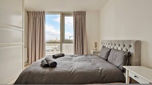 Ein Schlafzimmer mit einem großen Bett, einem Nachttisch, einer Lampe und Blick auf die Stadt durch das Fenster.