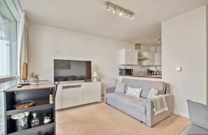 A modern living room with a grey sofa, a flat-screen TV, a coffee table, a bookshelf, and a kitchen area in the background. - Delightful Apartment Wandsworth (London)