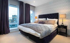 Een slaapkamer met een groot bed, een raam met gordijnen, een behang met patroon en een nachtkastje met een lamp.