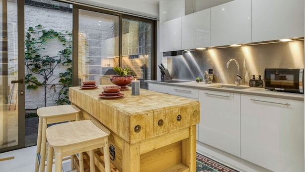 A modern kitchen with white cabinets, a wooden island, and a large window overlooking a brick wall with greenery. - Zen Chelsea Townhouse 3BR 3 5BA Terrace (London)
