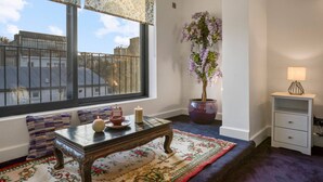 A room with a large window, a potted plant, a small table with candles, a bench with cushions, and a nightstand with a lamp. - Zen Chelsea Townhouse 3BR 3 5BA Terrace (London)
