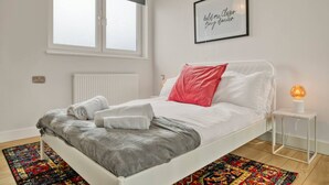Un dormitorio con cama, ventana, radiador, una cita enmarcada en la pared y una lámpara de noche.