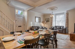 Ein Essbereich mit einem Holztisch, gedeckt für sechs Personen, eine Treppe und ein Wohnzimmer mit grauem Sofa und Fernseher.