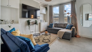 A modern living room with a sofa, a television, and a large window with curtains. - Chic 1bed w Patio Battersea (London)