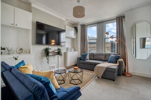 A modern living room with a sofa, a television, and a large window with curtains. - Chic 1bed w Patio Battersea (London)