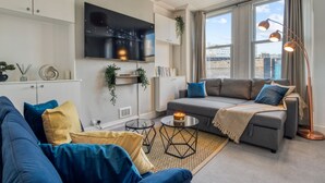 A modern living room with a grey sofa, blue cushions, a TV mounted on the wall, and a large window with curtains. - Chic 1bed w Patio Battersea (London)