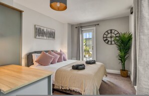 A bedroom with a bed, a bedside table, a wall clock, a window, and a potted plant. - Chic 1bed w Patio Battersea (London)