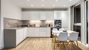 A modern kitchen with white cabinetry, a central island, and a dining area with a white table and chairs. - Modern 2 Bedroom Flat (London)