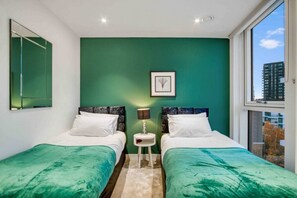 A room with two beds, a green accent wall, a framed picture, a small table with a lamp, and a window with a view of buildings. - Modern 2 Bedroom Flat (London)