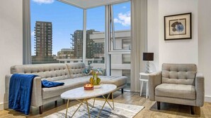 A modern living room with a sofa, armchair, coffee table, and a view of tall buildings. - Modern 2 Bedroom Flat (London)