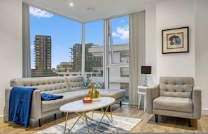 A modern living room with a sofa, armchair, coffee table, and a view of tall buildings. - Modern 2 Bedroom Flat (London)