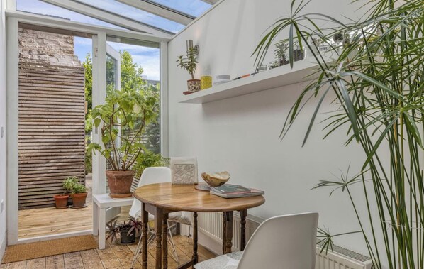 Une véranda dotée d’un plancher en bois, d’une table ronde entourée de chaises, de plantes en pot et offrant une vue sur la verdure environnante.
