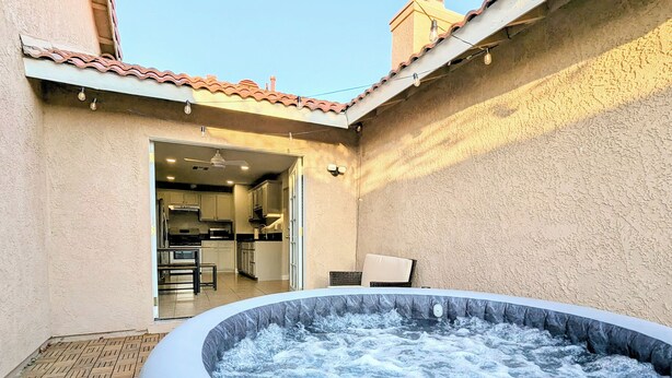 Un espace extérieur avec un jacuzzi, doté d’un sol carrelé, une cuisine visible à travers une porte ouverte et une chaise placée près du jacuzzi.