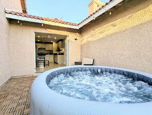 Un espace extérieur avec un jacuzzi, doté d’un sol carrelé, une cuisine visible à travers une porte ouverte et une chaise placée près du jacuzzi.