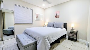 Une chambre à coucher comprenant un lit, des tables de chevet, un banc, un ventilateur de plafond, une fenêtre avec des stores et un miroir fixé au mur.