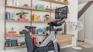 Et hjemmegymnasium med en motionscykel, et vægmonteret tv og en reol fyldt med bøger og pynteting.