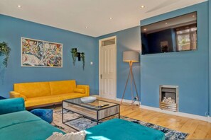 A living room with a yellow sofa, teal armchairs, a glass coffee table, a painting, a lamp, and a fireplace. - 107 Stonhouse Street Lovely 3-bed Clapham Common (London)
