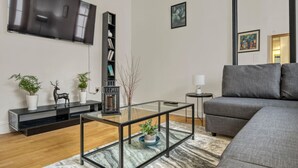 Ett modernt vardagsrum med en platt-TV, en bokhylla, ett soffbord i glas, en grå soffa och en mönstrad matta.