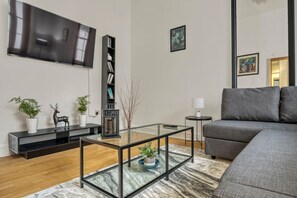 A modern living room with a flat-screen TV, a bookshelf, a glass coffee table, a grey sofa, and a patterned rug. - 2BR Retreat - Paddington Station (London)