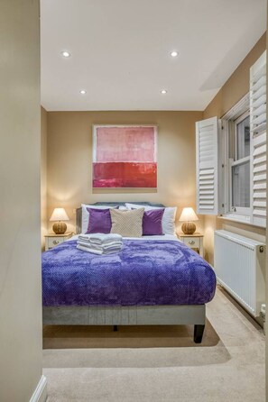 Een slaapkamer met een bed, twee nachtkastjes, een raam met luiken, een radiator en een schilderij aan de muur.