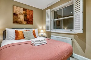 A neatly made bed with pillows and a blanket, a bedside lamp, a window with shutters, and a wall art piece. - Notting Hill Hyde Park - Cosy 2 Bed 2 Bath (London)