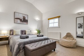 Een slaapkamer met een groot bed, nachtkastjes, een raam, een radiator, een poef en een schilderij aan de muur.
