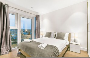 Ein modernes Hotelzimmer mit einem großen Bett, einem Nachttisch, einer Lampe und Blick ins Freie durch eine Schiebetür.