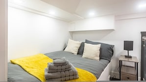 A bedroom with a bed, a bedside table with a lamp, a yellow blanket, and a stack of towels. - Quirky Studio in Central Reading (Reading)