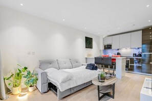 Uma sala de estar moderna com um sofá, uma mesinha com uma planta e uma área de cozinha com armários e eletrodomésticos.