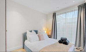 A bedroom with a bed, a bedside lamp, a window with curtains, and a view of the outside. - Cosy Manhattan Studio w Balcony (London)
