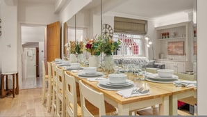 Zona pranzo con tavolo in legno apparecchiato per quattro, circondato da sedie bianche. Un grande specchio riflette l'ambiente, e un orologio è visibile sulla parete.