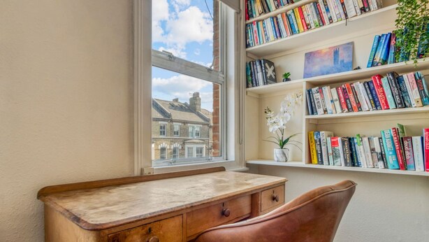 A desk with drawers, a chair, a bookshelf with books, a window with a view of buildings, and a small plant on the shelf. - 3bed Battersea Rise Books Charm and Style (London)