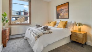 A bedroom with a large window, a bed with pillows, a bedside table with a lamp, and a radiator. - 3bed Battersea Rise Books Charm and Style (London)