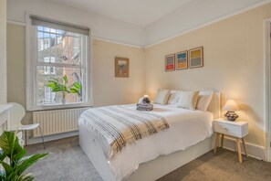 A bedroom with a large window, a radiator, a bedside table with a lamp, and a bed with a striped blanket. - 3bed Battersea Rise Books Charm and Style (London)