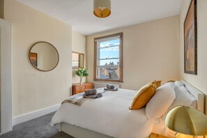 A bedroom with a bed, a round mirror, a window with a view, a dresser, and a lamp. - 3bed Battersea Rise Books Charm and Style (London)