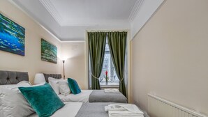 Camera d'albergo con due letti, una finestra con tende verdi, una lampada e opere d'arte alle pareti.